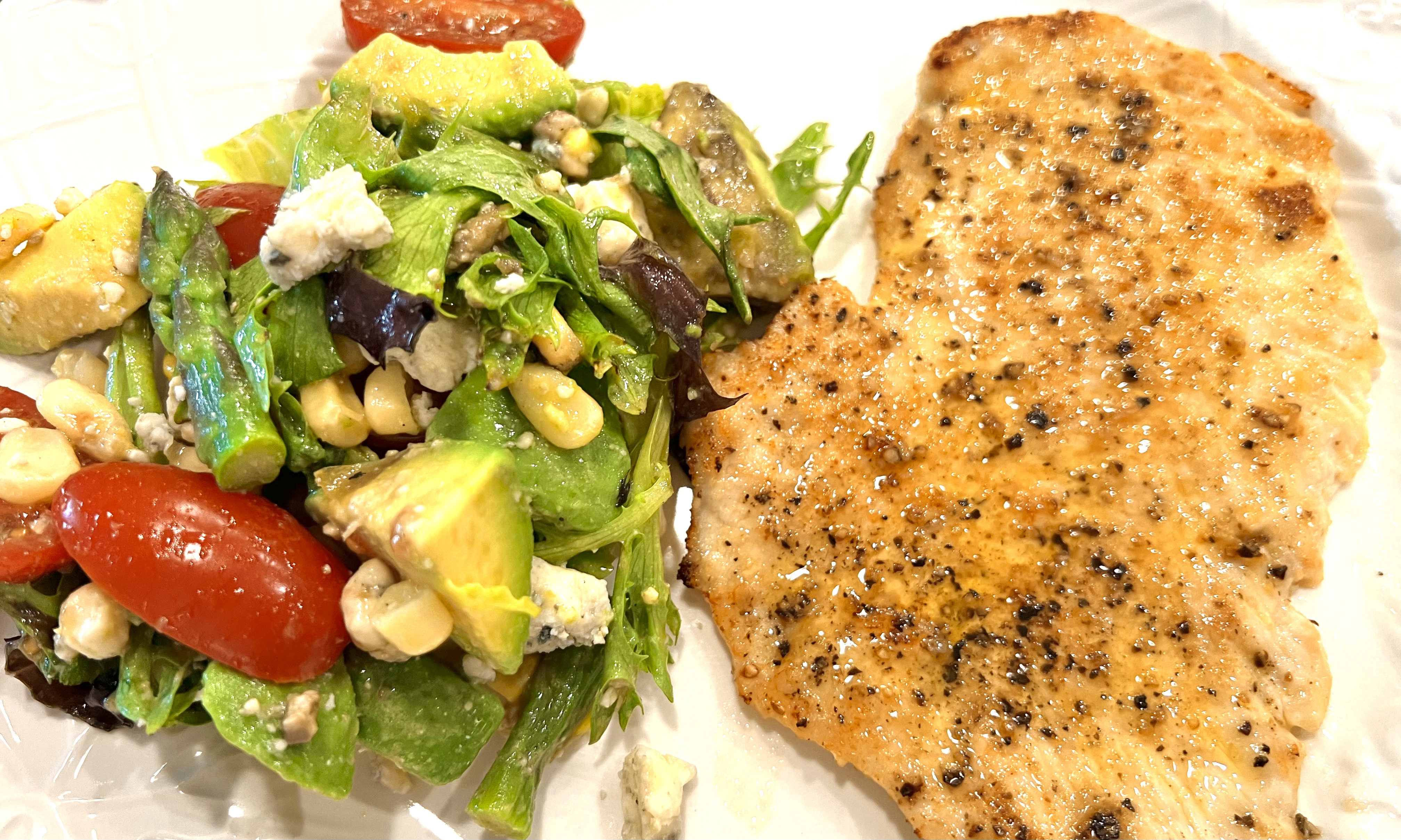 A colorful chopped chickpea and asparagus salad mixed with bright yellow sweet corn, halved red cherry tomatoes, diced green avocado, and chunks of crumbled Gorgonzola cheese, lightly drizzled with a clear lemon vinaigrette dressing served with chicken cutlet 